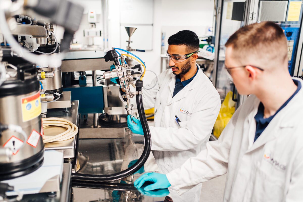 Two workers in lab coats and protective goggles using equipment